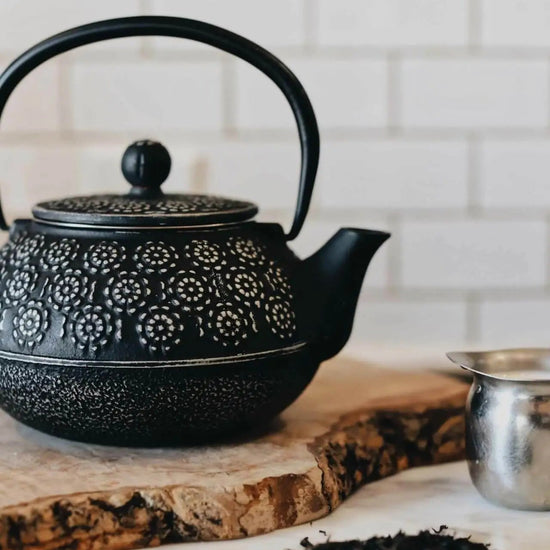 Black cast iron teapot on a wooden board with loose tea leaves on a marble surface.